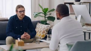 Man, patient or talking to psychologist on sofa in mental health, depression or anxiety counselling therapy. Psychology, stress or wellness counseling therapy help for person in rehabilitation clinic